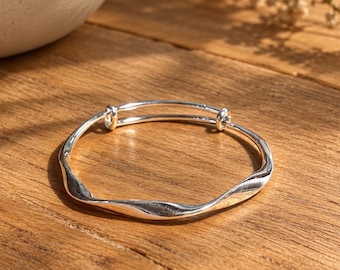 Könnte beinhalten: Ein silberfarbener, verstellbarer Armreif mit einem durchgehenden, wellenförmigen Metalldesign. Das Armband verfügt über zwei kleine kreisförmige Schlaufen an der Stelle, an der sich die Enden überlappen, wodurch der Durchmesser für einen bequemen Sitz angepasst werden kann.