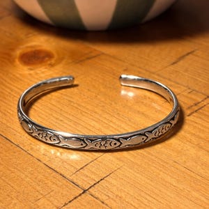 Puede incluir: Una pulsera de plata con un diseño repetido de peces. La pulsera abierta descansa sobre una superficie de madera. Los peces están estilizados y adornan la banda. Circunferencia aproximada de 17 cm.