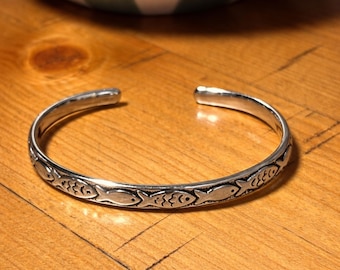 May include: A silver cuff bracelet featuring a repeating fish design. The open bracelet rests on a wooden surface. The fish are stylized and decorate the band. The bracelet is approximately 17 cm in circumference.