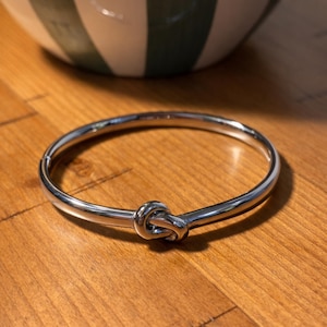 May include: A silver-colored bracelet with a knot design. The bracelet is circular and sits on a wooden surface. A white and green striped bowl is in the background.