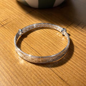 Könnte beinhalten: Ein verstellbares silbernes Armreifarmband mit Sternmuster. Das Armband besteht aus glänzendem Metall und liegt auf einer Holzoberfläche. Das Armband ist so konzipiert, dass es an verschiedene Handgelenkgrößen angepasst werden kann.