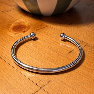 Könnte beinhalten: Ein glänzendes silbernes Armreif-Armband mit einer Kugel an jedem offenen Ende. Das Armband liegt auf einer Holzoberfläche. Eine weiß-grün gestreifte Schüssel befindet sich im Hintergrund.