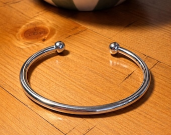 May include: A polished silver cuff bracelet with a ball at each end. The bracelet sits on a wooden surface. A white and green striped bowl is in the background.