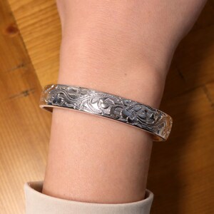 May include: A silver bangle bracelet with an engraved floral pattern. The bracelet, worn on a wrist, has a polished finish. The intricate design features swirling lines and stylized flower shapes, creating a decorative accessory. The bracelet's circumference is approximately 18 cm.