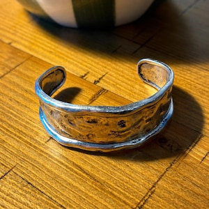 May include: A silver-coloured cuff bracelet with a textured surface. The bracelet has a hammered appearance and a wide band design. The edges are smooth and polished, creating a contrast with the rougher centre.