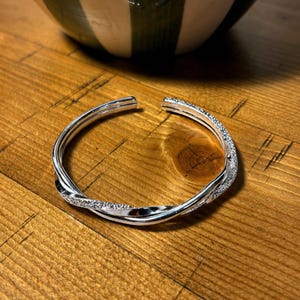 May include: A silver open cuff bracelet with a twisted design. One side is encrusted with small, sparkling stones. The bracelet sits on a wooden surface. The bracelet's diameter is approximately 6 cm.