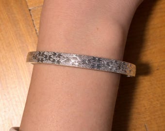 Może przedstawiać: Srebrna bransoletka z powtarzającym się wzorem rozbłysków gwiazd. Bransoletka ma styl mankietowy, a wzór jest wytrawiony w metalu. Bransoletka jest noszona na nadgarstku.