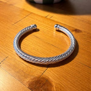 May include: A silver-colored, open cuff bracelet with a twisted rope design. The bracelet has rounded, smooth ends. The jewelry is displayed on a wooden surface.