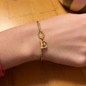 Op de afbeelding: Goudkleurige armband met de letter "E" als bedel. De armband heeft een delicaat ontwerp met een knoopdetail en een bungelende initiaal. De armband wordt om de pols gedragen.