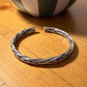 Può includere: Un bracciale a polsino in argento con un design intrecciato. Il bracciale è aperto alle estremità e poggia su una superficie di legno. Il bracciale ha un motivo strutturato e intrecciato ed è un gioiello.