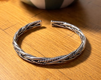 May include: A silver cuff bracelet with a braided design. The bracelet is open at the ends and sits on a wooden surface. The bracelet has a textured, woven pattern and is a piece of jewellery.