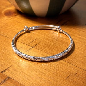 Può includere: Un braccialetto a cerchio regolabile argentato con un design strutturato e fantasia. Il braccialetto è realizzato in metallo lucido e ha uno stile semplice ed elegante. Il braccialetto è mostrato su una superficie di legno.
