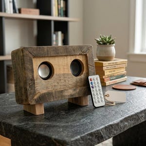 May include: A rectangular wooden speaker featuring a rustic, weathered brown frame with two circular speaker drivers, one silver and one black. The speaker stands on two light-colored wooden feet and is accompanied by a small white remote control.