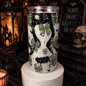 May include: A cylindrical glass tumbler featuring a white and black wavy pattern decorated with black and white striped bows, neon green slime accents, spider webs, and small black bats, topped with a clear plastic lid.