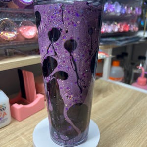 May include: A clear double-walled tumbler filled with a purple liquid, dark black swirling ink-like patterns, and fine iridescent glitter. The cup features a transparent lid and a cylindrical shape, showcasing a fluid motion effect inside the walls.