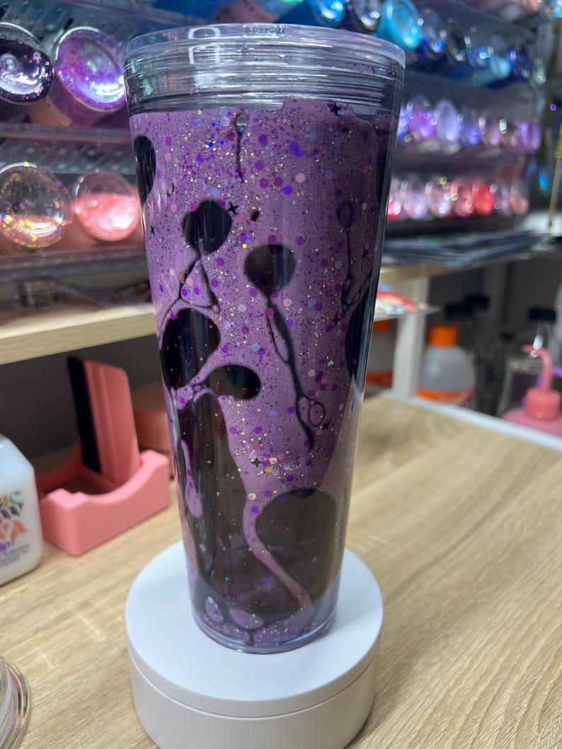 May include: A clear double-walled tumbler filled with a purple liquid, dark black swirling ink-like patterns, and fine iridescent glitter. The cup features a transparent lid and a cylindrical shape, showcasing a fluid motion effect inside the walls.