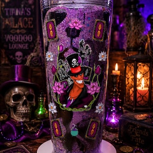 May include: A clear plastic tumbler filled with purple glitter liquid, featuring a graphic of a man in a black top hat with a skull and crossbones, surrounded by pink lotus flowers, tarot cards with hand symbols, and a small green cauldron charm.