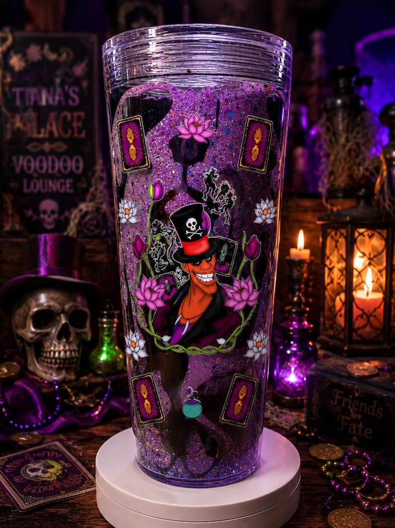 May include: A clear plastic tumbler filled with purple glitter liquid, featuring a graphic of a man in a black top hat with a skull and crossbones, surrounded by pink lotus flowers, tarot cards with hand symbols, and a small green cauldron charm.