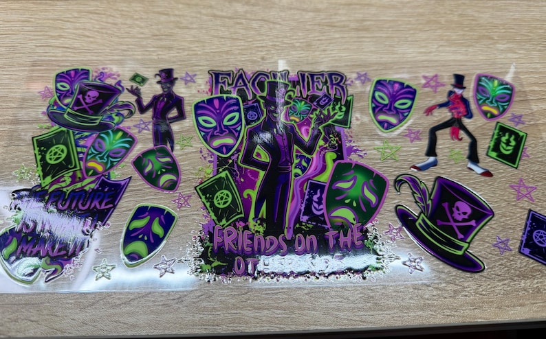 May include: A clear transfer sheet featuring a purple and green graphic design with a central figure in a top hat, surrounded by theatrical masks, playing cards, and skull-adorned hats. The text reads "FACILIER" and "FRIENDS ON THE OTHER SIDE".
