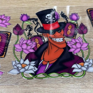 May include: A clear transfer sheet featuring a central character wearing a black top hat with a skull and crossbones, surrounded by purple and white lotus flowers and several rectangular tarot cards displaying a golden hand symbol on a purple background.