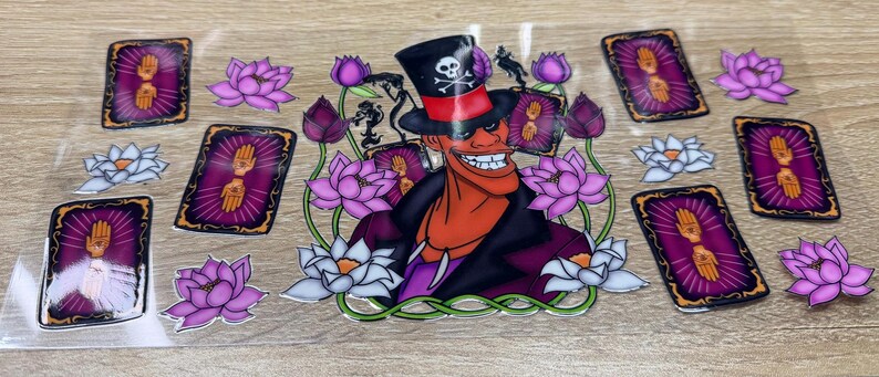 May include: A clear transfer sheet featuring a central character wearing a black top hat with a skull and crossbones, surrounded by purple and white lotus flowers and several rectangular tarot cards displaying a golden hand symbol on a purple background.
