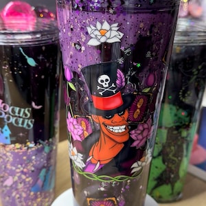 May include: A purple glitter tumbler with a light purple straw and a rhinestone-encrusted lid. The cup features a graphic of a character in a top hat with a skull and crossbones, surrounded by white and pink lotus flowers and tarot cards.