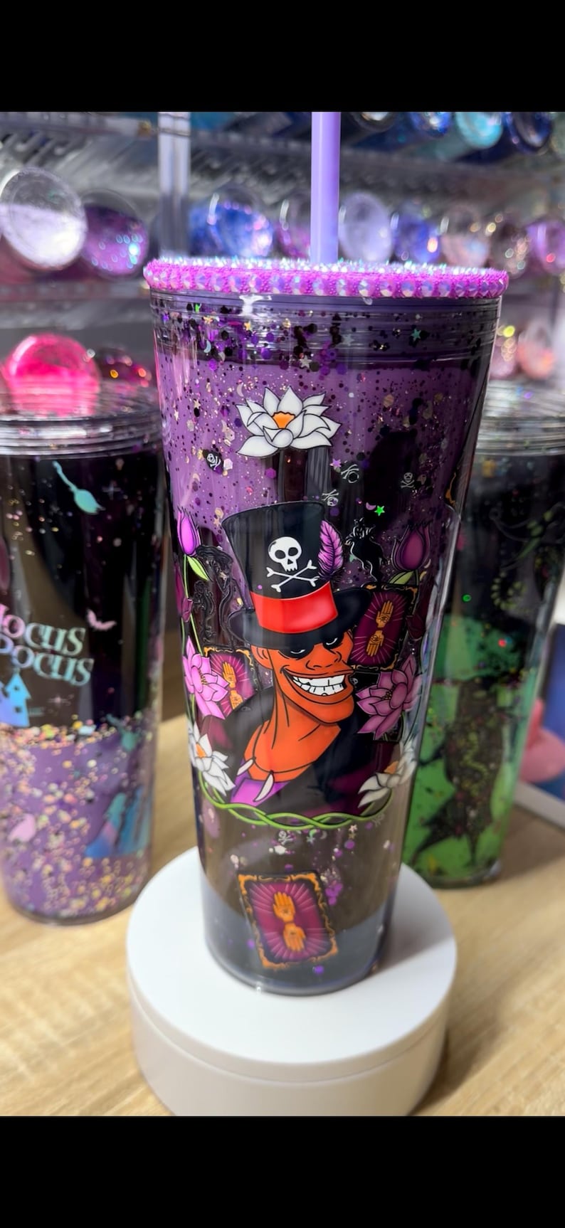 May include: A purple glitter tumbler with a light purple straw and a rhinestone-encrusted lid. The cup features a graphic of a character in a top hat with a skull and crossbones, surrounded by white and pink lotus flowers and tarot cards.