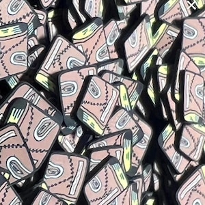 May include: A collection of small, rectangular, tan-colored book-shaped charms. Each charm features a black stitched line across the cover, a white eye graphic, and yellow page accents. The items are arranged in a dense, overlapping pile.