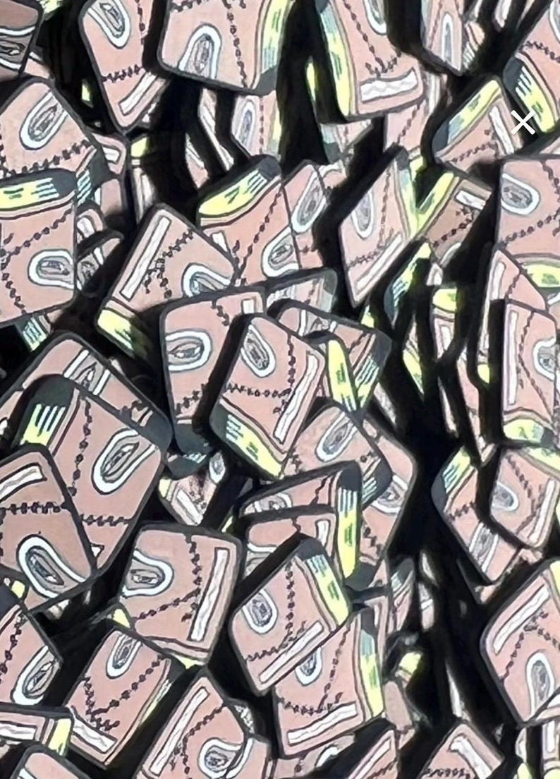 May include: A collection of small, rectangular, tan-colored book-shaped charms. Each charm features a black stitched line across the cover, a white eye graphic, and yellow page accents. The items are arranged in a dense, overlapping pile.