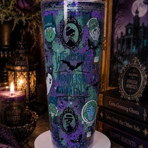May include: A clear plastic tumbler filled with purple and green glitter, featuring printed graphics of ghosts, ornate frames, bats, and the text Welcome Foolish Mortals in a stylized blue font against a swirling purple and green background.