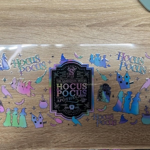 May include: A clear sticker sheet featuring iridescent Hocus Pocus themed designs, including silhouettes of the Sanderson Sisters, cauldrons, broomsticks, bats, and cats. The central label reads The Sanderson Sisters Hocus Pocus Apothecary.