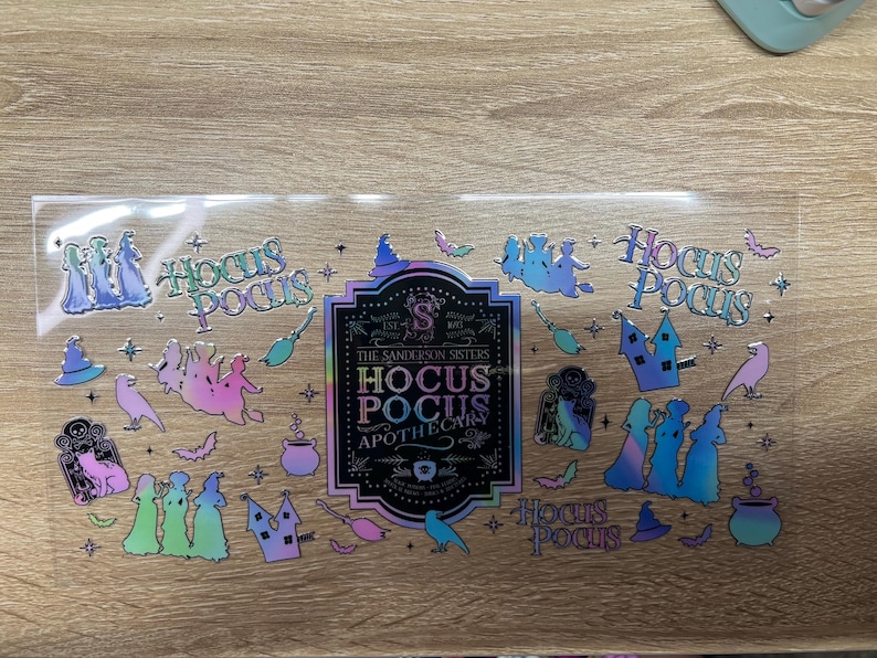 May include: A clear sticker sheet featuring iridescent Hocus Pocus themed designs, including silhouettes of the Sanderson Sisters, cauldrons, broomsticks, bats, and cats. The central label reads The Sanderson Sisters Hocus Pocus Apothecary.