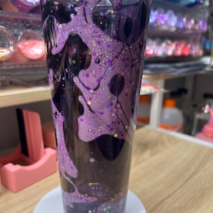 May include: A clear plastic tumbler featuring a double-walled design filled with a dark liquid and a thick, viscous purple substance that drips down the interior walls. The purple layer is embedded with various sizes of purple and gold glitter.