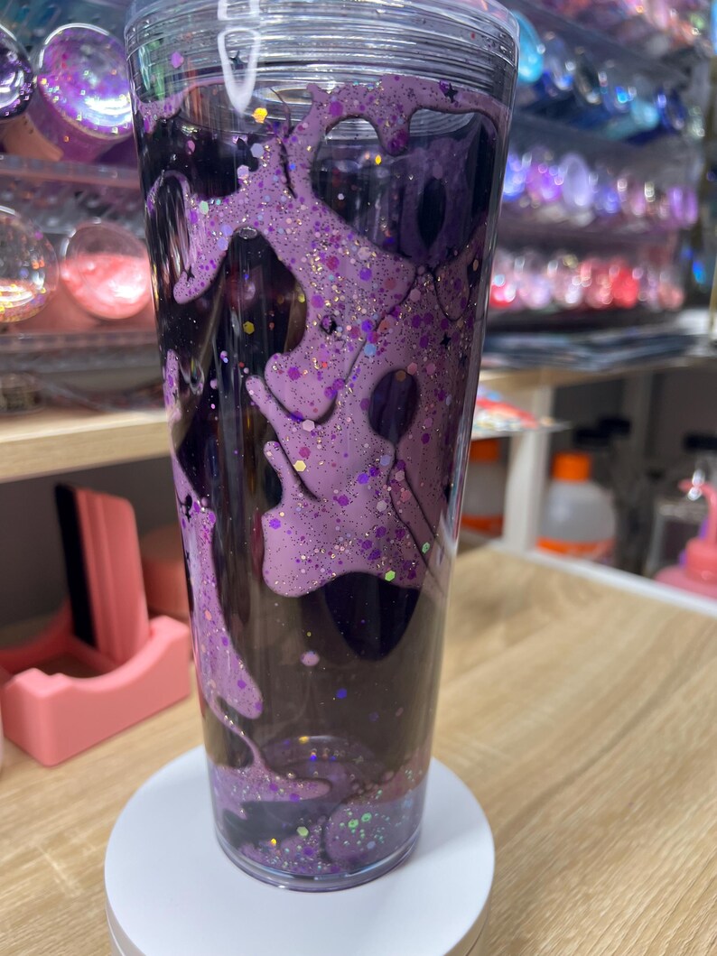 May include: A clear plastic tumbler featuring a double-walled design filled with a dark liquid and a thick, viscous purple substance that drips down the interior walls. The purple layer is embedded with various sizes of purple and gold glitter.