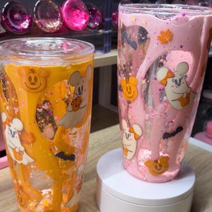 May include: Two clear tumblers with lids, one filled with orange liquid and the other with pink liquid. Both feature internal drip designs, glitter, and various Halloween-themed decals including ghosts, pumpkins, and bats with mouse-like ears.