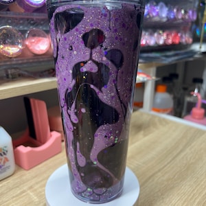 May include: A clear double-walled tumbler featuring a purple glitter liquid filling that forms the silhouette of a dog face. The design includes dark purple accents, star-shaped glitter pieces, and a transparent lid, creating a layered effect.