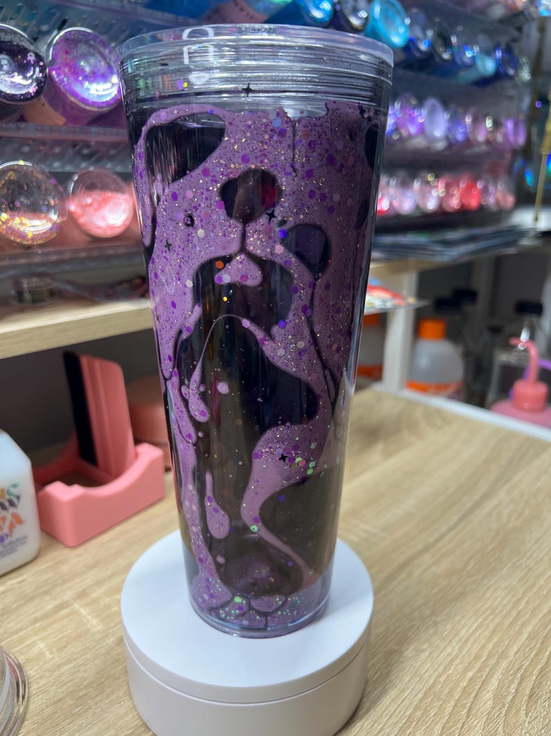 May include: A clear double-walled tumbler featuring a purple glitter liquid filling that forms the silhouette of a dog face. The design includes dark purple accents, star-shaped glitter pieces, and a transparent lid, creating a layered effect.