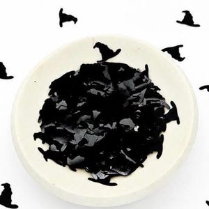 May include: A small, round, off-white ceramic bowl filled with numerous black, flat, witch hat-shaped confetti pieces. Additional black witch hat confetti pieces are scattered around the bowl on a plain white surface.