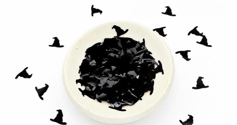 May include: A small, round, off-white ceramic bowl filled with numerous black, flat, witch hat-shaped confetti pieces. Additional black witch hat confetti pieces are scattered around the bowl on a plain white surface.