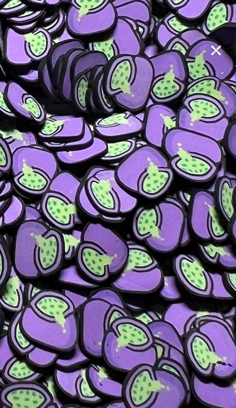 May include: A collection of small, flat, purple-colored charms shaped like halved fruits. Each piece features a light green interior with dark green seeds and a black outline, creating a uniform, repeating pattern of identical fruit-themed items.