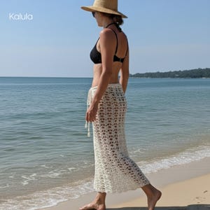 May include: A woman wearing a black bikini top and a cream-colored crochet skirt stands on a sandy beach. She is wearing a straw hat and sunglasses. The skirt is long and has a delicate, open weave design. The word "Kalula" is in the top left corner.