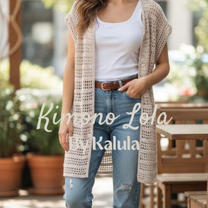 May include: A beige, open-weave crochet kimono-style cardigan with the text "Kimono Lola by Kalula". The long, sleeveless cardigan is worn over a white vest top and blue jeans. A brown belt is visible.