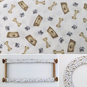 May include: White fabric featuring a repeating pattern of tan dog bones, food bowls, and grey paw prints. The fabric is stretched over a wooden embroidery hoop. Ideal for pet-themed crafts and home decor projects.