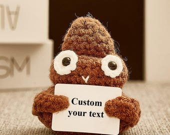 May include: A brown crocheted plush toy shaped like a swirl, featuring two large white-rimmed eyes with black pupils and a small white stitched mouth. The plush holds a rectangular white sign with the black text Custom your text.