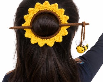 May include: A crochet hair accessory featuring a yellow sunflower ring with a brown centre, secured by a brown stick. A small, crocheted bumblebee with white wings and yellow and black stripes hangs from the end of the stick by a brown cord.