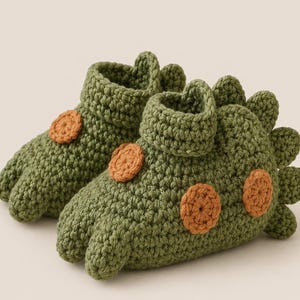 May include: A pair of olive green crochet booties shaped like dinosaurs. Each bootie features a textured surface, a folded cuff at the ankle, and small orange circular patches along the side. A row of green spikes runs along the back heel.