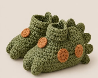 May include: A pair of olive green crochet booties shaped like dinosaurs. Each bootie features a textured surface, a folded cuff at the ankle, and small orange circular patches along the side. A row of green spikes runs along the back heel.