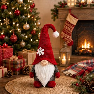 May include: A crochet gnome figurine featuring a tall, pointed red hat with a white snowflake applique and a white pom-pom at the tip. The gnome has a beige nose, a long white beard, a gray body, red mittens, and red feet, standing upright.