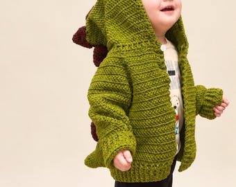 May include: A green crocheted hooded cardigan featuring a dinosaur-inspired design with dark brown spikes along the back and hood, a ribbed hem and cuffs, and a single button closure at the neck, worn over a graphic t-shirt and black trousers.