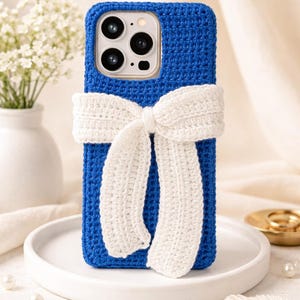 May include: A blue crochet phone case with a white crochet bow. The phone case is a solid blue colour, and the bow is a contrasting white. The case is designed to fit a smartphone.